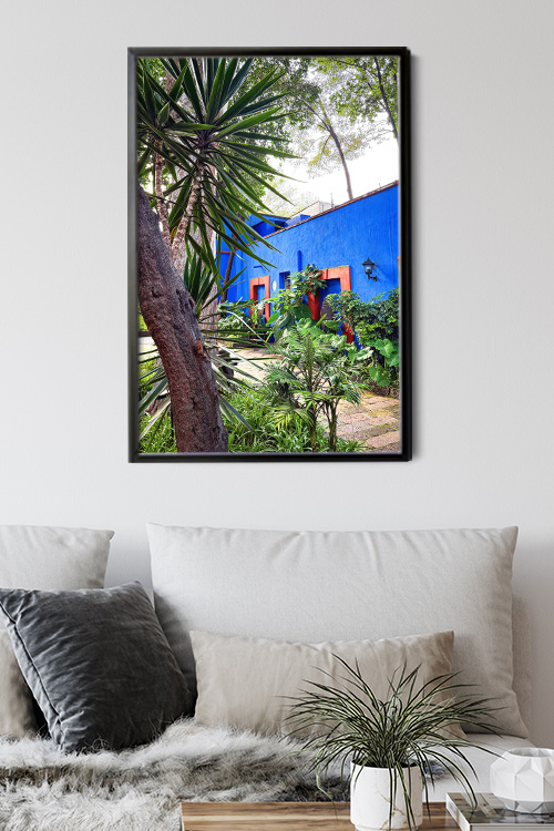 A framed poster print hanged on a white wall showing an image of trees outside of a blue building